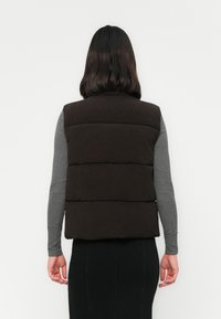 Kvinne med skulderlangt mørkt hår som har på seg en svart polstret vest over en grå langermet skjorte og et svart skjørt, vendt bort fra kameraet på en hvit bakgrunn.