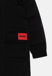 Svart puffjakke med glatt overflate, som har en fremtredende rød "HUGO" logo patch på venstre lomme og en borrelås-lukking på ermet.