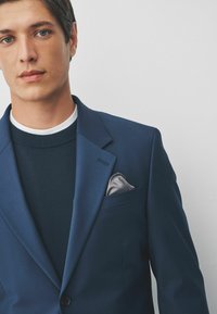 Marineblauwe blazer met een enkele knoopsluiting, scherpe revers en een grijze zakdoek. Daaronder een donker marineblauw sweater met een witte kraag.