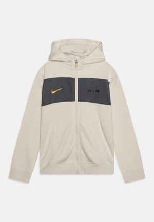 Jasnobeżowa bluza z kapturem zapinana na zamek, z szarym panelem na piersi, złoto-czarnym logo Nike oraz przednimi kieszeniami. Gładka faktura tkaniny.