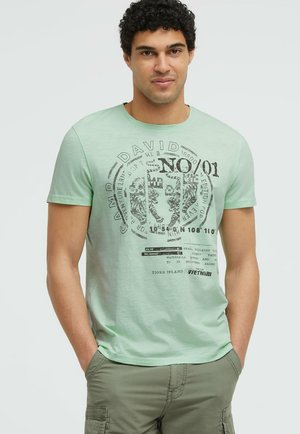 Mann mit lockigem Haar, der ein hellgrünes T-Shirt mit Grafik und olivfarbene Cargohosen trägt, steht mit den Händen in den Taschen.