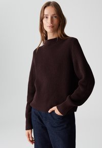 Pull en maille côtelée marron avec un col montant et des manches larges, associé à un pantalon en denim foncé. Texture douce et coupe décontractée.