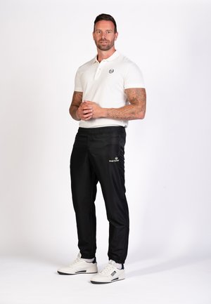 Witte polo met kraag, zwarte sportbroek met logo-detail en witte sneakers met marineblauwe accenten. Glad stof, casual pasvorm.