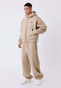 Felpa beige con zip e ampie tasche frontali, abbinata a pantaloni larghi coordinati. Il tessuto ha una texture liscia; il modello indossa sneakers bianche.