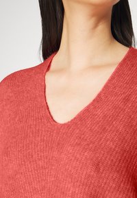 Röd stickad tröja med V-ringning, texturerad tyg och avslappnad passform. Accentuerad med råa kanter för en avslappnad look.