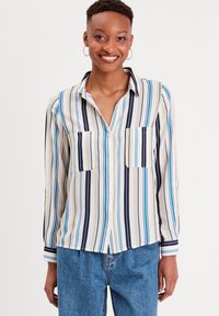 Blouse rayée beige, bleue et marine ; à boutons avec des manches longues et deux poches poitrine ; tissu léger au toucher agréable.