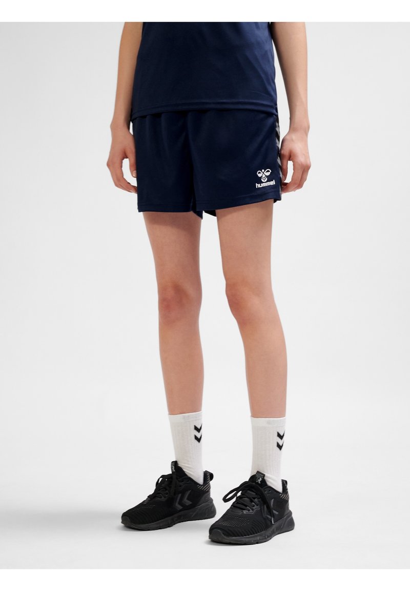 Personne portant un short de sport bleu marine et des baskets noires avec des chaussettes blanches, debout devant un fond clair uni.