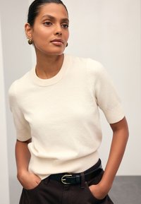 Pull crème à manches courtes avec un col et des poignets côtelés. Associé à un pantalon foncé et une ceinture noire avec une boucle dorée.