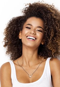 Collana d'argento con pendente circolare, indossata sopra una canotta bianca, completata da capelli ricci che incorniciano il viso. Sorriso luminoso e caloroso visibile.