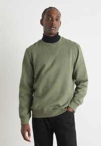 Olíva zöld crewneck pulóver puha pamutból, ribbelt mandzsettákkal és aljjal. Kicsi hímzett logóval a mellrészen. Fekete garbó fölé viselve.