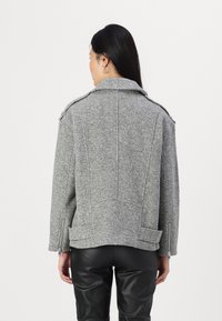 Veste en mélange de laine grise avec un col structuré, des coutures visibles, des zips sur les côtés et une coupe décontractée et oversize. Portée sur un pantalon noir.