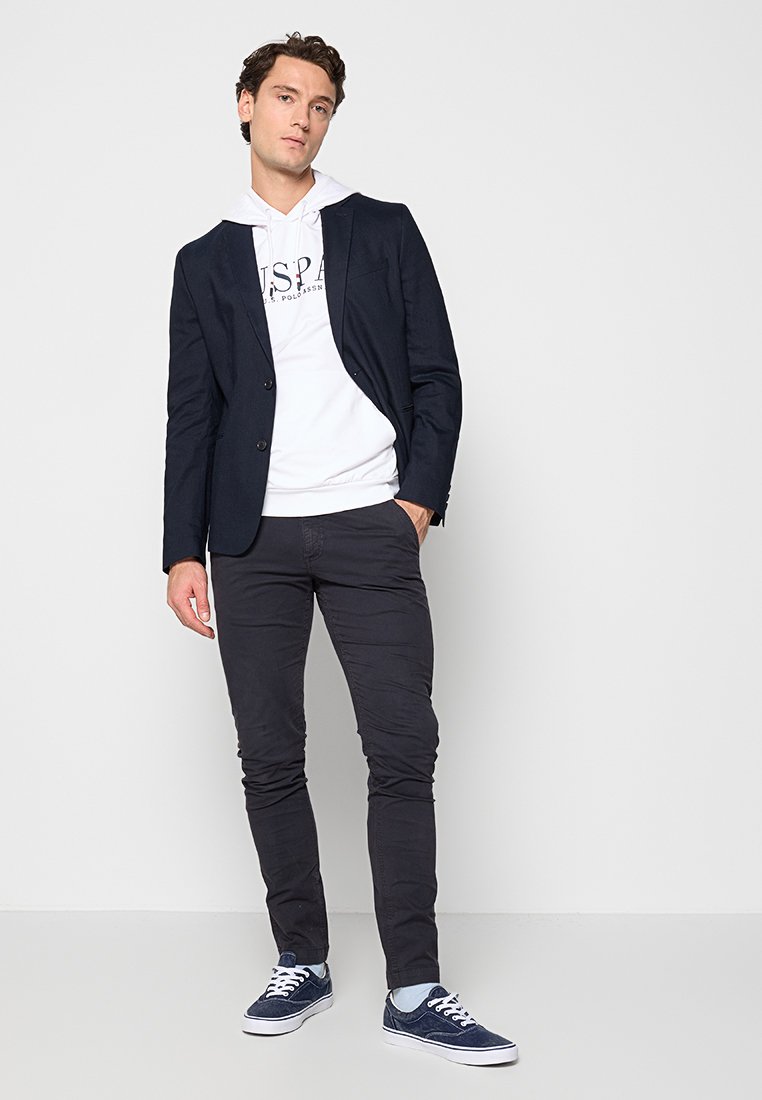 Blazer navy sobre una sudadera blanca, pantalones oscuros y zapatos navy con suela blanca. Diseño ajustado, textura suave, con un logo en la sudadera.