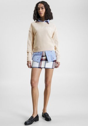 Felpa beige con logo "HILFIGER", sovrapposta a una camicia a righe blu e bianche, abbinata a una mini gonna a fantasia e mocassini neri.