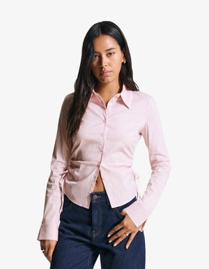 Light pink button-up shirt with long sleeves, striped texture, side drawstring details, and a fitted design paired with blue jeans.