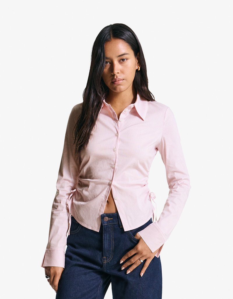 Camicia button-up rosa chiaro con maniche lunghe, texture a righe, dettagli con cordoncino laterale e un design aderente abbinata a jeans blu.