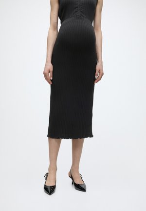 Schwangere Frau trägt ein schwarzes ärmelloses, geripptes Kleid und schwarze Lack-Slingback-Pumps und steht vor einem schlichten weißen Hintergrund.