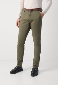 Pantalon chino vert olive à coupe slim, avec poches latérales et ceinture en cuir marron. Des mocassins noirs complètent le look.