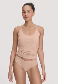 Vrouw in bijpassende beige cami-top met kant en ondergoed, staand tegen een effen lichte achtergrond.