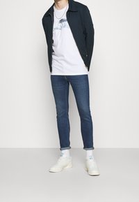 Navy jacket, white graphic tee, slim-fit blue jeans, and white sneakers with rolled cuffs; casual outfit with contrasting colors and textures.
