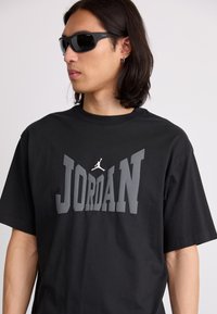 Juoda medvilninė marškinėlių su ryškiu pilku "JORDAN" spaudiniu ir Jumpman logotipu. Modelis dėvi akinius nuo saulės, marškiniai turi trumpas rankoves ir klasikinį kirpimą.
