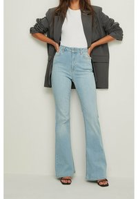 Jeans a vita alta a zampa d'elefante di un blu chiaro con una texture denim liscia, tasche frontali e una silhouette aderente, abbinati a una maglietta bianca e a un blazer grigio.
