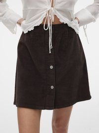 Jupe mini en velours côtelé marron avec des boutons sur le devant et un ourlet légèrement évasé. Tissu texturé avec des lignes verticales. Associée avec un haut blanc.