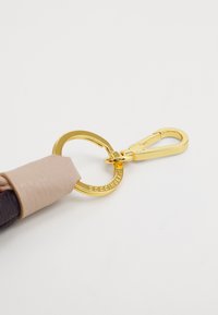 Chaveiro de couro com um anel e fecho de metal dourado, apresentando um detalhe em couro rosa em uma das extremidades. Texto suave e formato circular.