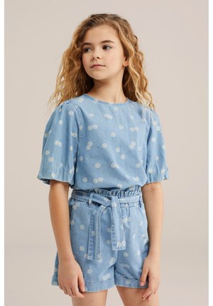 Hajlított hajú lány, kék, margarétás mintás rövid ujjú felsőt és hozzá illő, öves sortot visel, oldalra néz.