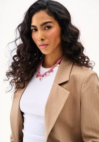 Žena s kučeravými tmavými vlasmi, oblečená v béžovom sako s tenkými pruhmi, bielom topu a červenom kockovanom náhrdelníku s príveskami v tvare srdca.