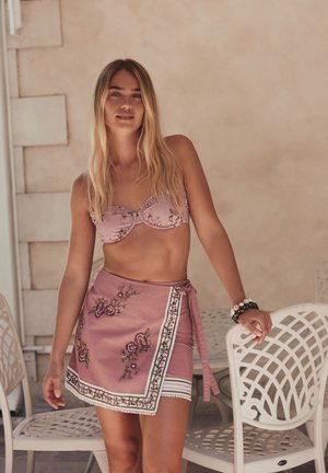 Mladá žena s dlouhými blond vlasy, která má na sobě růžové květované bikini horní díl a odpovídající zavinovací sukni, stojí vedle bílých židlí s mřížkovým vzorem uvnitř.