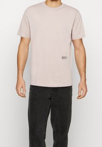 Camiseta de algodón rosa claro con mangas cortas y cuello redondo, que presenta un pequeño logotipo negro de REPLAY "Second Life" en el lado izquierdo.