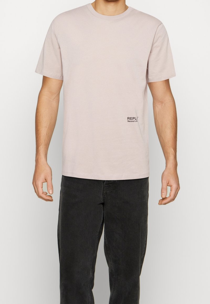 Camiseta de algodón rosa claro con mangas cortas y cuello redondo, que presenta un pequeño logotipo negro de REPLAY "Second Life" en el lado izquierdo.