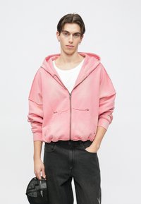 Rosa hoodie med dragkedja, tillverkad av mjukt tyg med en avslappnad passform, utrustad med stor huva, framfickor och slitna kanter. Kombinerad med mörka jeans.
