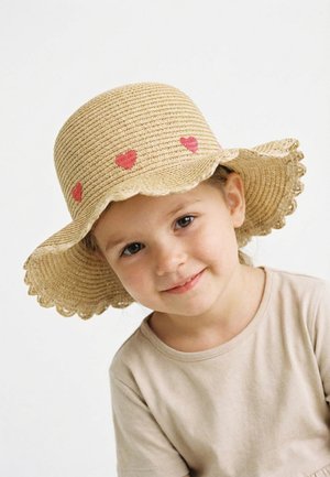 Junges Kind trägt einen beigefarbenen breitkrempigen gewebten Hut mit roten Herzdekorationen und lächelt sanft vor einem einfarbigen weißen Hintergrund.