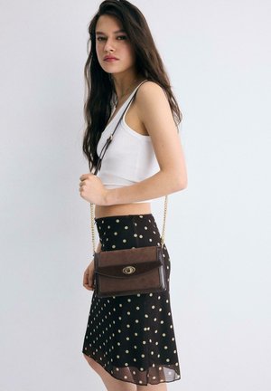 Mlada ženska v beli majici brez rokavov in črni krili z belimi pikami, ki nosi rjavo torbo čez ramo z zlatim verižnim trakom na enobarvnem ozadju.