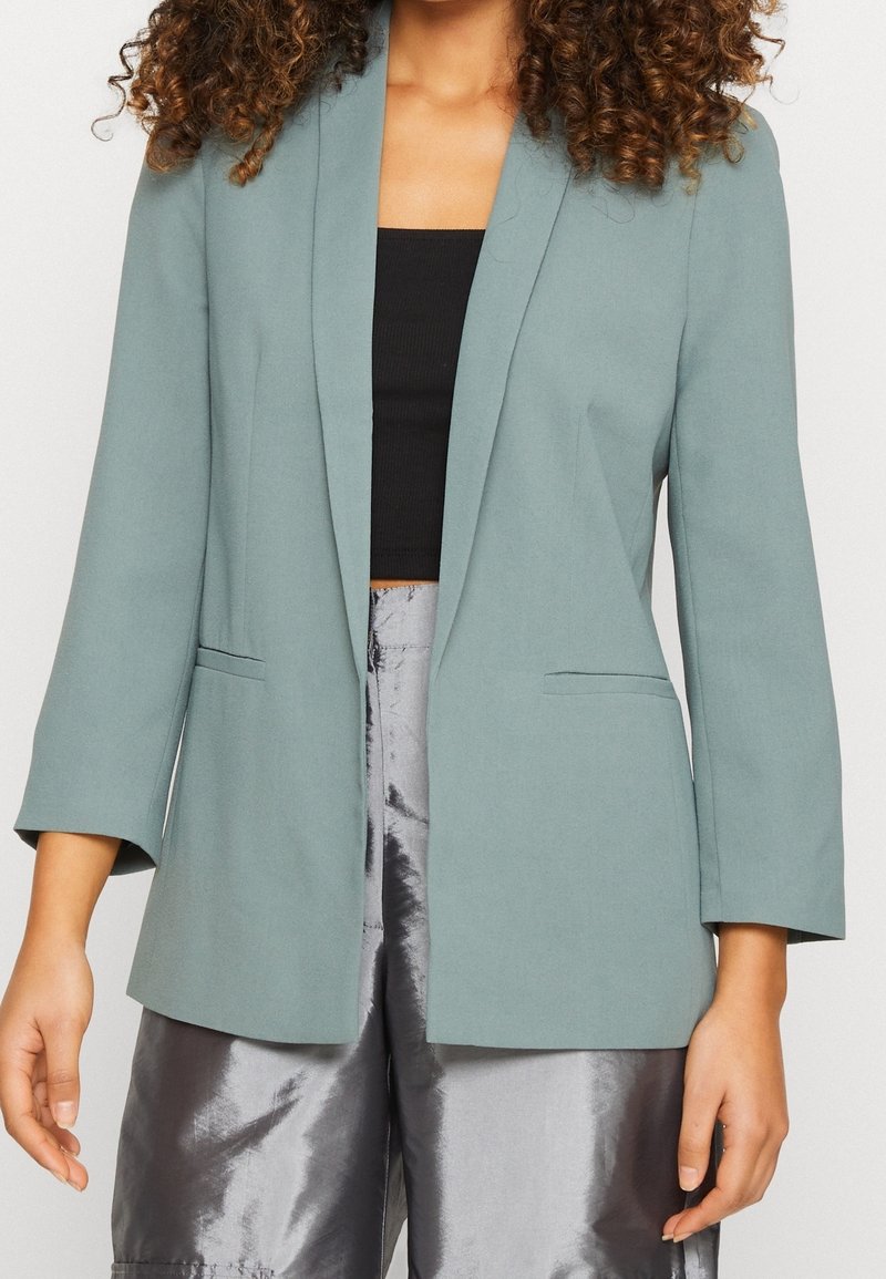 Femme portant un blazer vert clair à manches trois-quarts, un top court noir et un pantalon large argenté brillant.