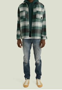 Man som bär en grön och vit rutig jacka över en mörkgrön huvtröja, urblekta blå jeans och beige sneakers med vita snören, stående mot en ljus bakgrund.