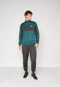 Teal og svart treningsjakke med glidelås, kombinert med grå bukser. Begge har et sporty design og logo på brystet og låret. svarte joggesko.