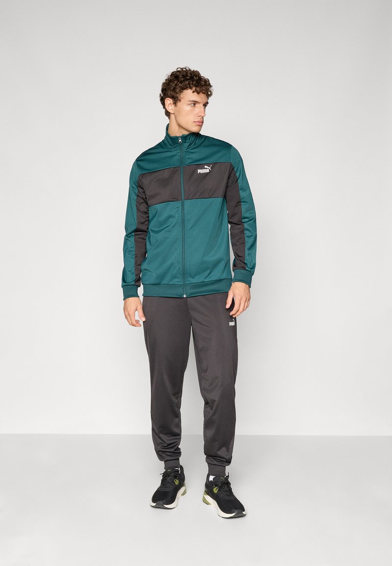 Teal og svart treningsjakke med glidelås, kombinert med grå bukser. Begge har et sporty design og logo på brystet og låret. svarte joggesko.