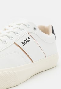 Sneaker in pelle bianca con una texture liscia, caratterizzata da una sottile striscia colorata tan, logo "BOSS" e una suola in gomma di colore chiaro.