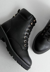 Stivali alla caviglia in pelle nera con occhielli in tono oro, design lace-up e suola nera robusta. Le cuciture dettagliate lungo i bordi aggiungono stabilità.