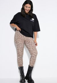 Žena v černém oversized tričku, béžových legínách s leopardím vzorem a černých robustních botách, usmívající se a pózující před bílým pozadím.