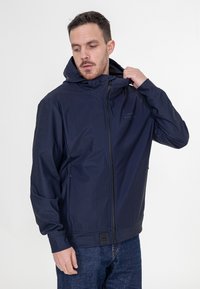 Granatowa wodoodporna kurtka z kapturem, z zamkiem z przodu, dwoma bocznymi kieszeniami i elastycznymi mankietami. Posiada subtelną markę na klatce piersiowej.