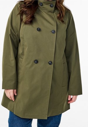 Frau trägt einen olivgrünen, doppelreihigen Mantel mit schwarzen Knöpfen und hohem Kragen, kombiniert mit blauen Jeans, vor weißem Hintergrund.