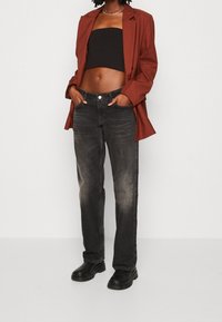 Femme portant un blazer rouille, un haut tube noir, un jean noir délavé et des bottes noires épaisses, debout les mains dans les poches.