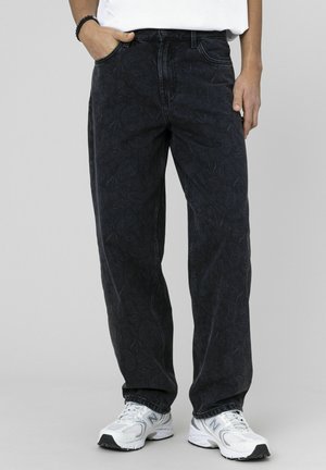 Jean en denim noir avec un motif texturé subtil, coupe droite, cinq poches et fermeture par bouton, porté avec des chaussures de sport blanches.