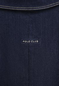 Mörkt denimtyg med en slät textur. Har en vit sömnadsdetalj och en rektangulär etikett med texten "POLO CLUB" i vitt.