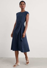 Robe bleu marine à pois blancs, manches courtes, détail de ceinture à la taille, et jupe évasée, accompagnée de sandales légères à lanières.