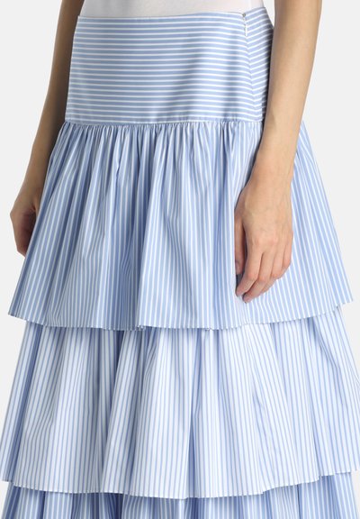 Blå og hvide lodrette stribede lagdelt nederdel med rynket stof, vist fra taljen til midt på låret med en hånd hvilende på siden.