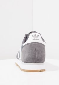 Rear view of a gray Adidas sneaker with white stripes and a gum sole on a white surface and background.